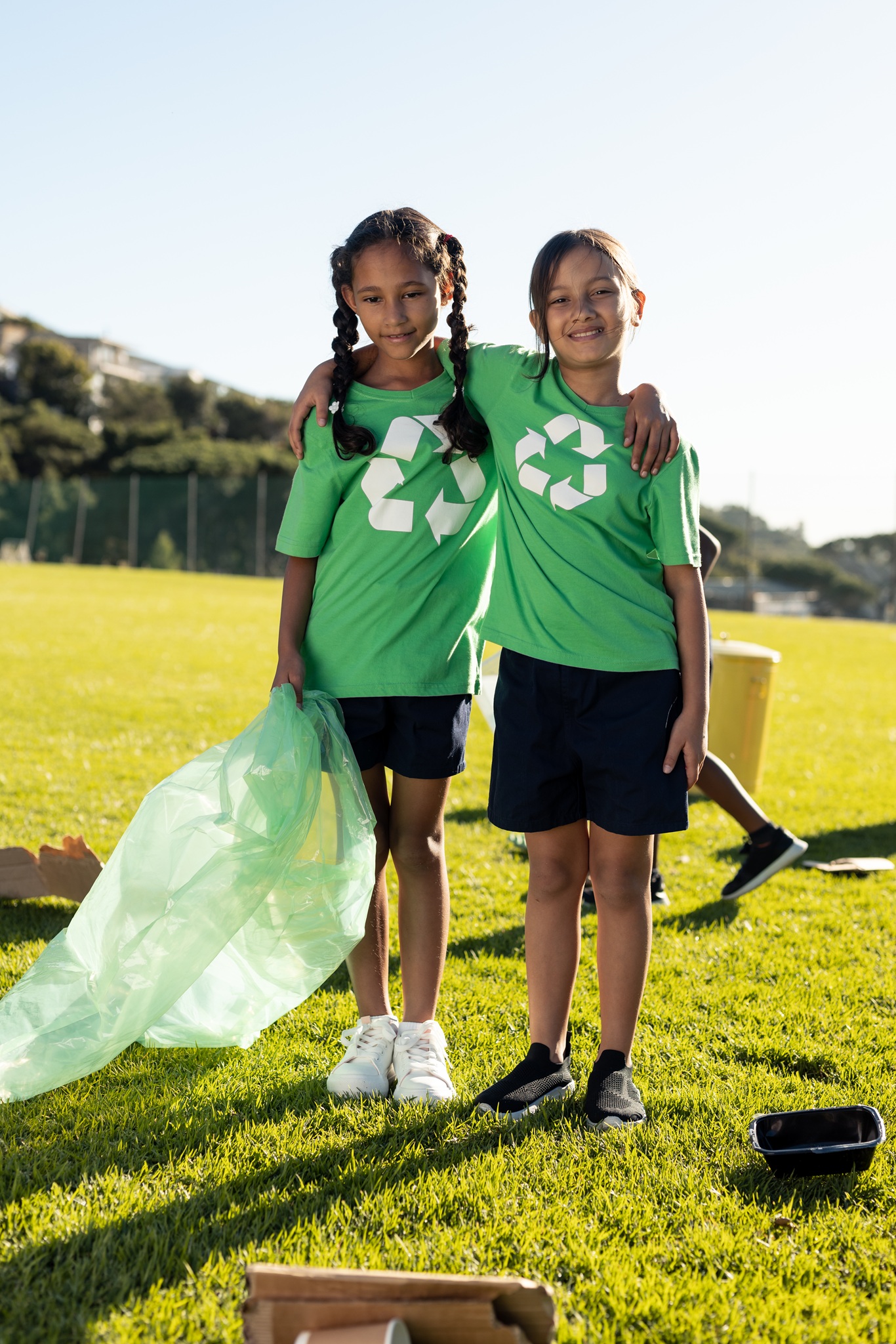 Portrait of happy biracial girls collecting recycling embracing in sunny elementary school field. Recycling, environment, education, childhood, development, learning and school, unaltered.
