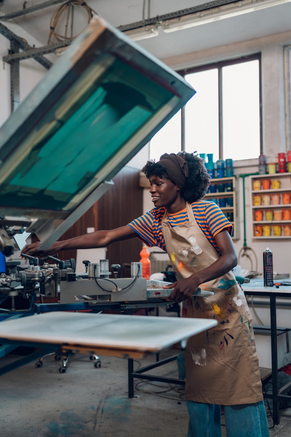 Young multicultural female graphic worker standing at printing workshop near silkscreen printing press and adjusting it for printing. Smiling diverse graphic expert preparing printing press for work.