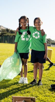 Portrait of happy biracial girls collecting recycling embracing in sunny elementary school field. Recycling, environment, education, childhood, development, learning and school, unaltered.