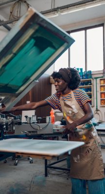 Young multicultural female graphic worker standing at printing workshop near silkscreen printing press and adjusting it for printing. Smiling diverse graphic expert preparing printing press for work.